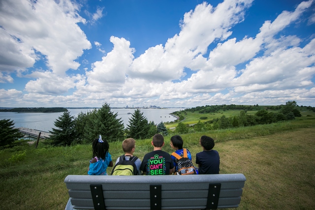 Group of children sit on a bench on a drumlin hill on Spectacle Island overlooking Boston Harbor
